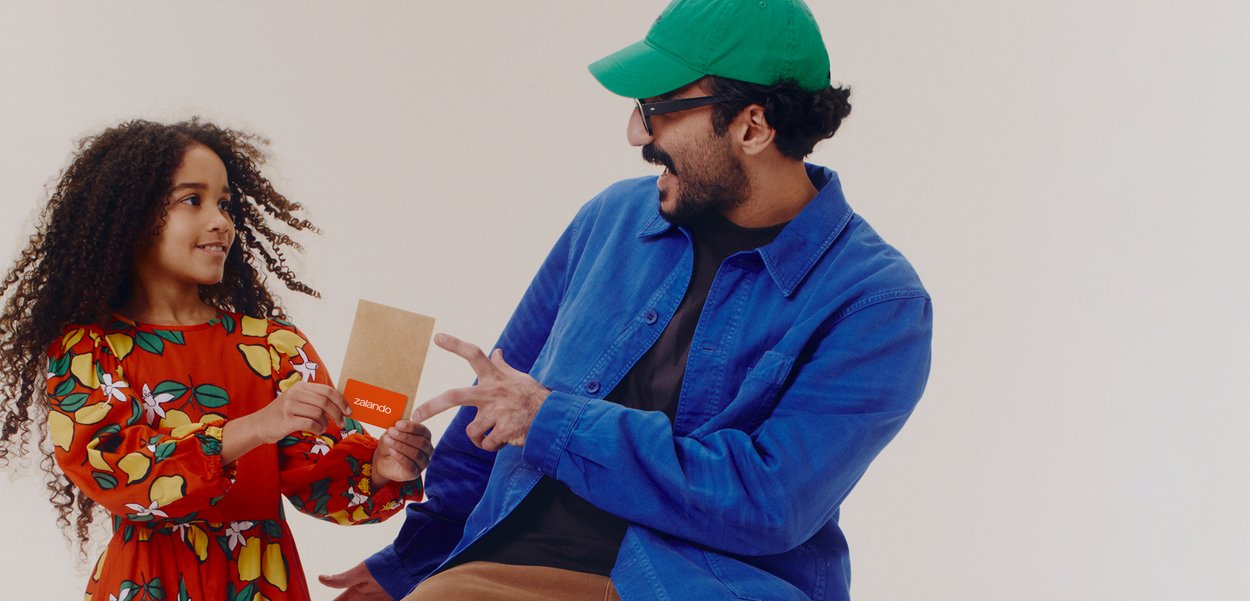 Ragazza in vestito floreale porge una carta arancione a un uomo con cappello verde e giacca blu, entrambi sorridenti e impegnati nella conversazione.