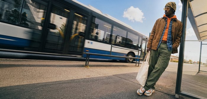 Un homme en vêtements décontractés et avec un foulard sur la tête se tient à un arrêt de bus, tenant un sac fourre-tout, tandis qu'un bus bleu et blanc passe par une journée ensoleillée.