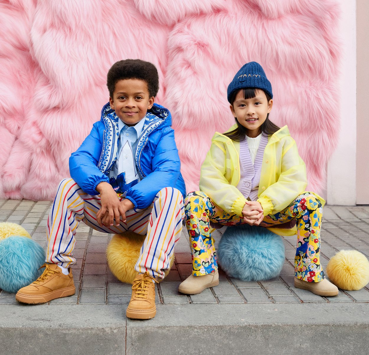 Un garçon en doudoune bleue éclatante, chemise blanche et pantalon rayé ; une fille en imperméable jaune et pantalon à motifs, tous deux assis sur des boules duveteuses.