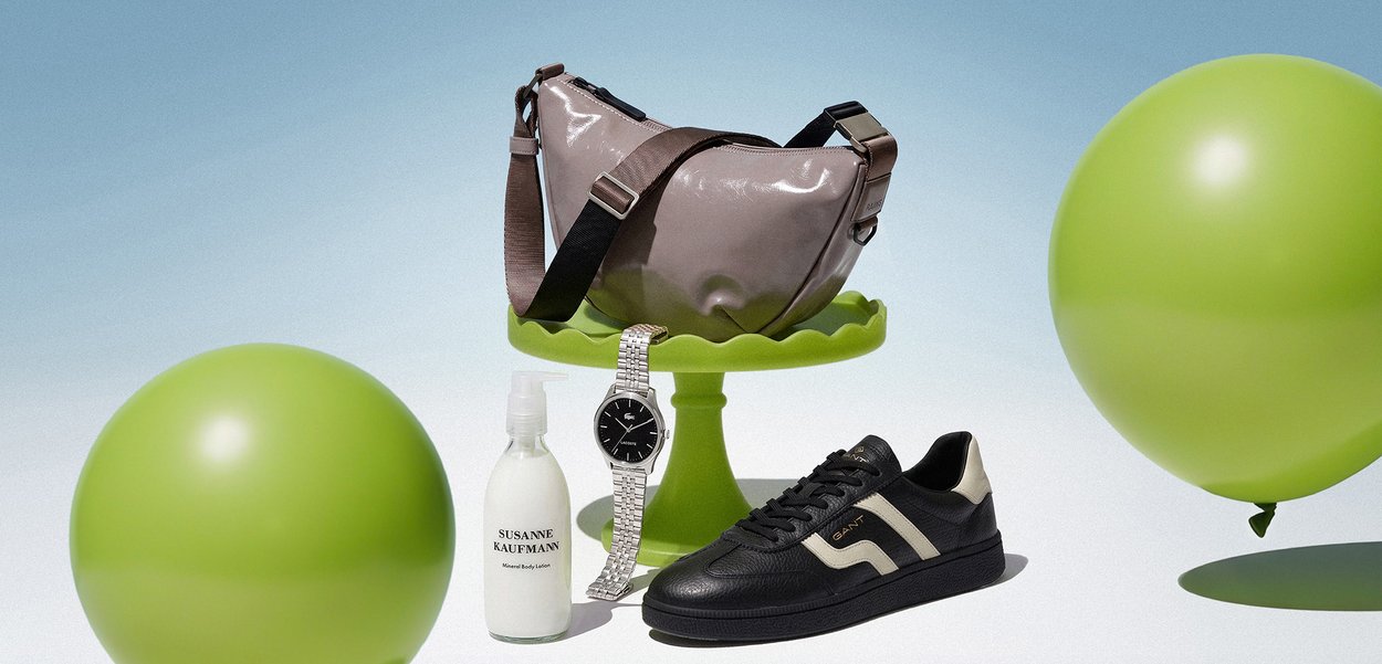Bolso bandolera de color púrpura claro sobre un soporte verde con una zapatilla negra y beige, reloj plateado, botella blanca de loción y dos globos verdes sobre fondo azul.