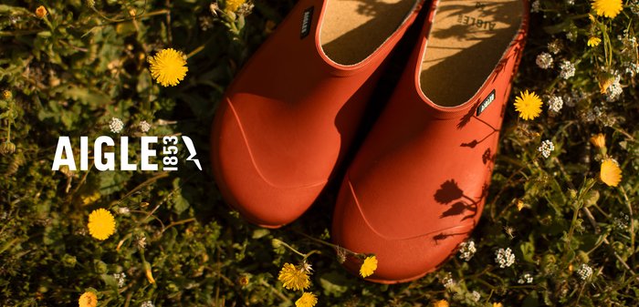 Sabots en caoutchouc rouge avec surface texturée et dessus ouvert, reposant sur de l'herbe verte parmi des fleurs sauvages jaunes et blanches. Logo visible.