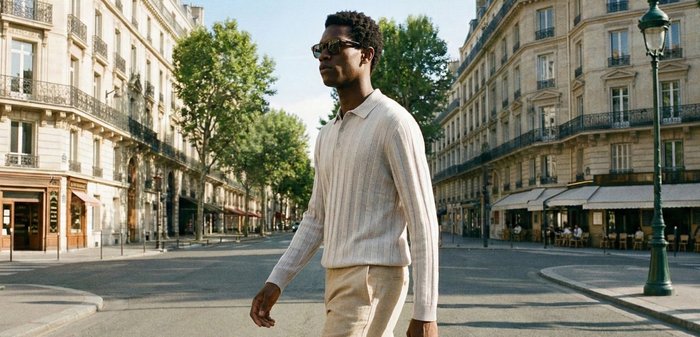 Homme portant des lunettes de soleil et un pull léger, marchant dans une rue de la ville bordée de bâtiments classiques européens et de cafés en plein air.