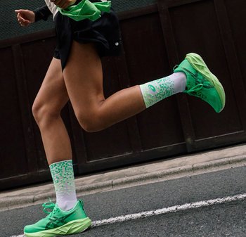 Benen van een hardloper in volle pas, met felgroene hardloopschoenen en witte sokken met groene patronen, op een stedelijke straat naast de stoep.