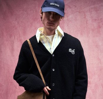 Giovane uomo che indossa un berretto blu navy, un cardigan nero con bottoni e logo, una camicia a righe gialla, e porta una borsa a tracolla marrone su sfondo rosa.