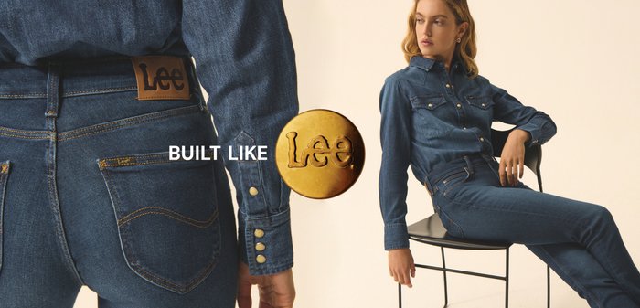 Femme assise sur une chaise noire portant une chemise en denim bleu foncé et un jean, regardant sur le côté, avec le logo "Built Like Lee" visible.