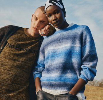 Maglione marrone lavorato a maglia con scritta "Nessun problema"; maglione lavorato a maglia con sfumatura blu; entrambi indossati sopra denim; copricapi a contrasto, ambiente naturale.