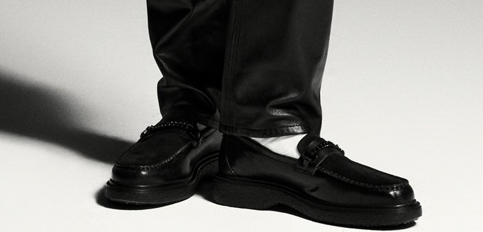 Pieds portant des mocassins en cuir noir avec détail chaîne et des chaussettes blanches, debout les jambes croisées sur un fond clair.