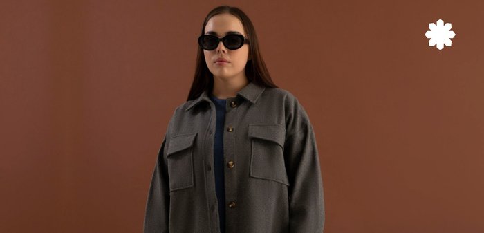 Haut en polaire grise à boutons avec deux poches avant, boutons dorés et coupe décontractée, porté par-dessus un pull bleu, sur un fond marron.