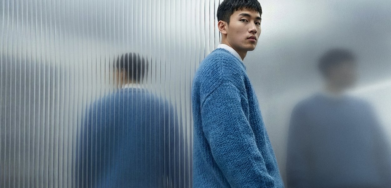 Νεαρός άνδρας με μπλε πλεκτό πουλόβερ στέκεται δίπλα σε έναν τοίχο από υαλοπίνακα με υφή, που αντανακλά δύο θολές εικόνες του.