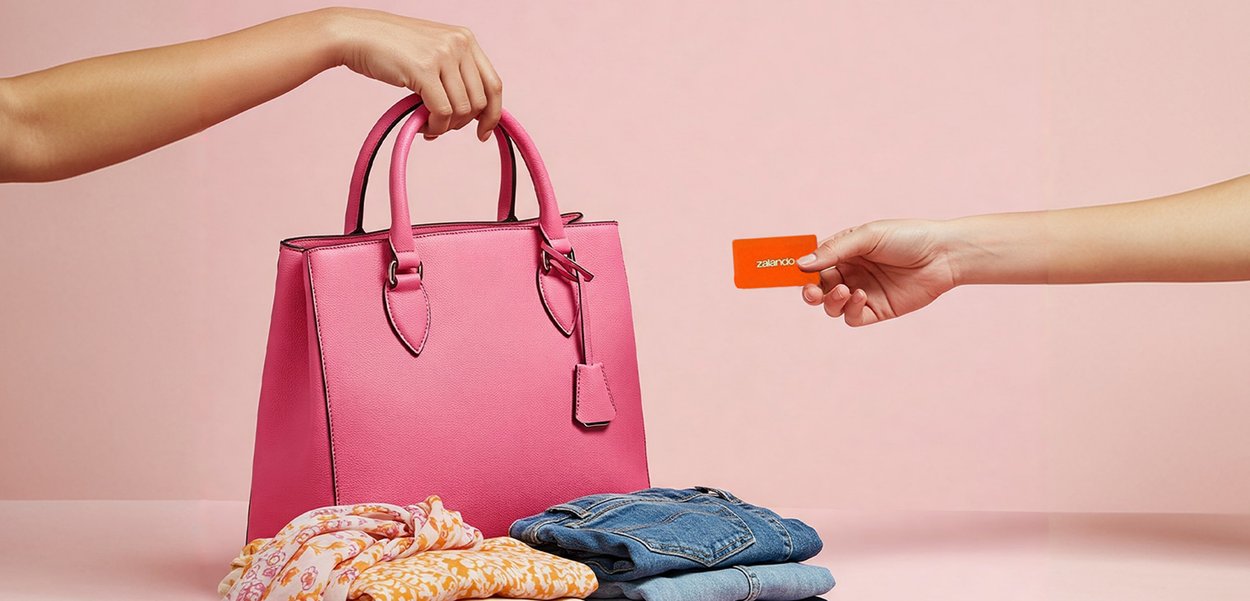Mano sosteniendo un bolso rosa junto a ropa doblada con otra mano ofreciendo una tarjeta naranja de Zalando sobre un fondo rosa.