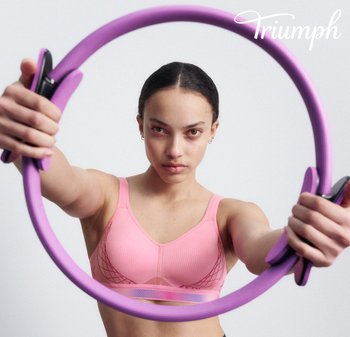 Lila Fitnessring, der mit beiden Händen gehalten wird und schwarze Akzente aufweist. Die Person trägt einen hellrosa Sport-BH mit strukturierter Stoffoberfläche und Netzdetails.