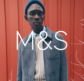 Szara zamszowa kurtka na kremowym swetrze w wzory i jasnoniebieskiej koszuli, dopełnionej niebieską dzianinową czapką, na czerwonym tle.