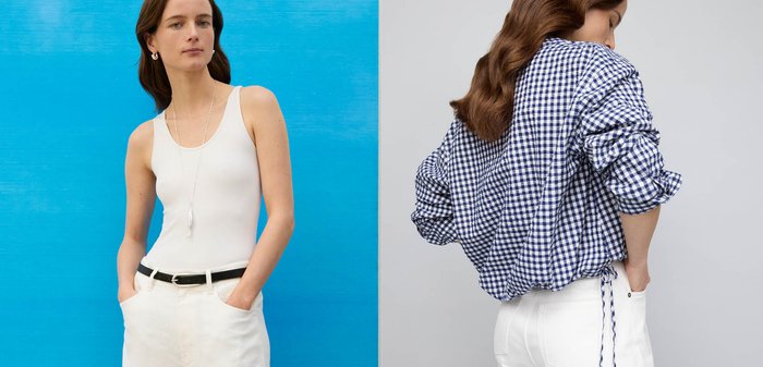 Femme aux longs cheveux bruns portant une chemise à carreaux bleue et blanche à manches longues et un pantalon blanc, debout avec une main dans la poche sur fond gris.