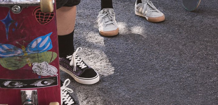 Three pairs of feet wearing sneakers stand near skateboards on asphalt, with partial skateboard deck visible in foreground.