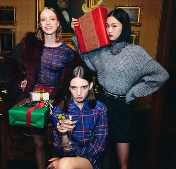 Trzy kobiety w pomieszczeniu z prezentami świątecznymi; jedna trzyma kieliszek koktajlowy, druga niesie duży zapakowany prezent na ramieniu, ubrane świątecznie.