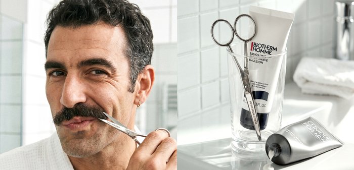 Un homme taille sa moustache avec des ciseaux à côté d'un lavabo de salle de bain en tenant des ciseaux de toilettage et des tubes de produits de soin pour la peau Biotherm Homme.