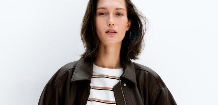 Femme portant une veste en cuir marron foncé sur un t-shirt blanc rayé, debout les mains dans les poches devant un fond blanc uni.