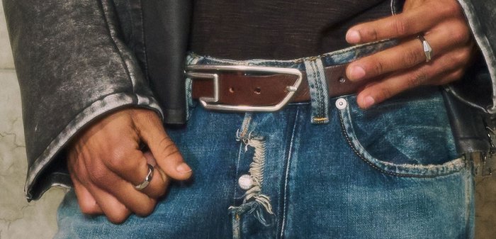 Hände mit Ringen, die einen braunen Gürtel an abgenutzten blauen Jeans anpassen, getragen mit einem schwarzen Hemd und einer grauen Lederjacke.