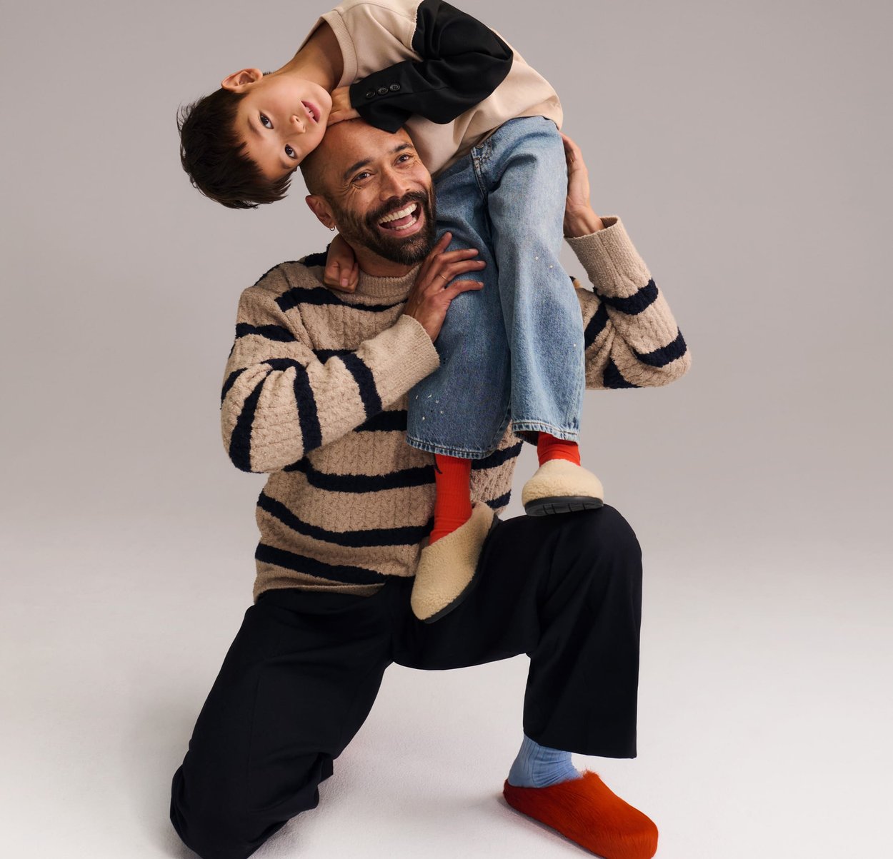 Bărbat în genunchi, zâmbind, ținând un copil cu capul în jos peste umăr, pe un fundal simplu.