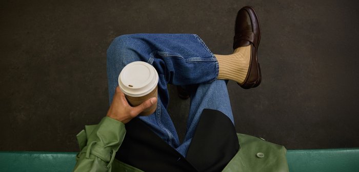 Personne portant des mocassins marron, des chaussettes beige, un jean bleu et un manteau vert tient une tasse de café à emporter, les jambes croisées sur un sol sombre.