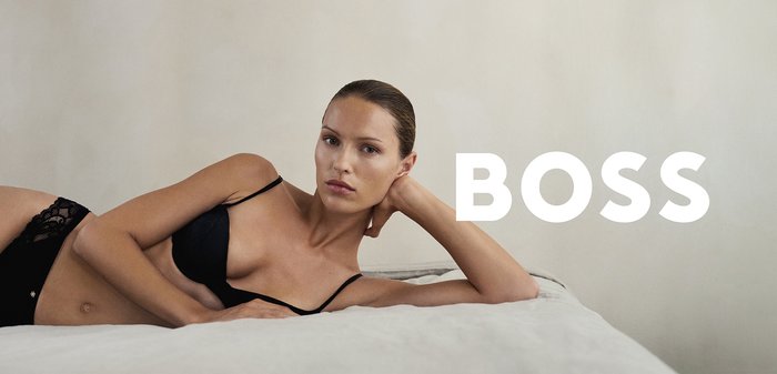 Woman reclining on a bed wearing a black bra, resting her head on her hand, with the word "BOSS" in bold white letters nearby.