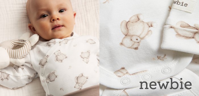 Bébé allongé portant un grenouillère blanc avec des imprimés d'ours en peluche beige et un pantalon marron, tenant un jouet doux rayé.