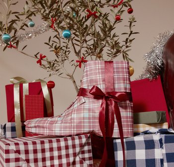 Czerwono-różowa w kratkę paczka w kształcie buta, z bordową wstążką w kształcie kokardy. Otoczona różnorodnie wzorzystymi zapakowanymi prezentami.