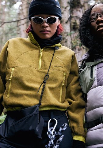 Ελιάς πράσινο fleece πουλόβερ με φερμουάρ στην τσέπη του στήθους και πλαϊνές τσέπες με φερμουάρ. Μαύρο παντελόνι με λευκή κορδέλα. Δυο άτομα σε ένα δασικό τοπίο.