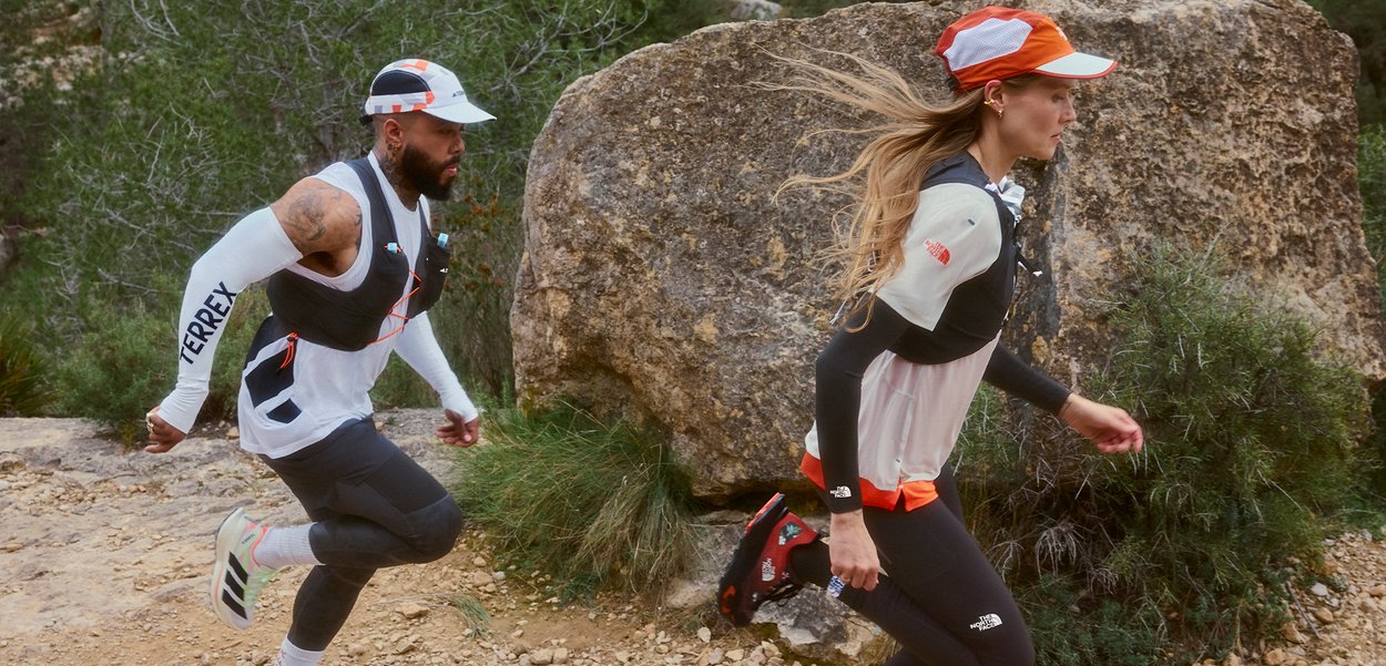 Un hombre y una mujer con ropa deportiva y gorras corren por un sendero rocoso de tierra, con vegetación y grandes rocas cercanas.