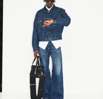 Persona con giacca di jeans scura e jeans a gamba larga, camicia bianca, occhiali da sole, che guarda l'orologio e tiene una borsa nera con tracolla larga.