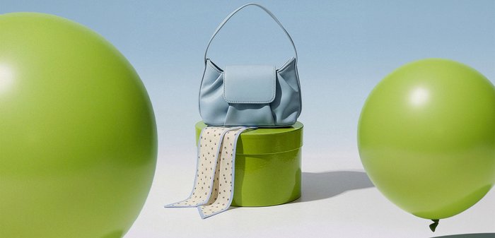 Svjetloplava torba na okrugloj limunasto zelenoj kutiji s maramom s točkicama preko nje, okružena s dvije velike zelene balone na svijetloj pozadini.