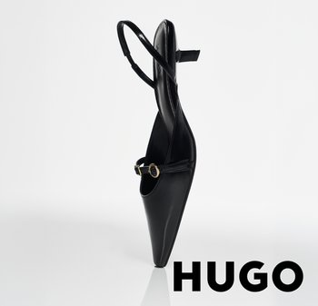 Zapato de tacón alto negro con punta puntiaguda, correa fina en el tobillo y pequeña hebilla, de pie sobre una superficie blanca reflectante, con la marca HUGO.
