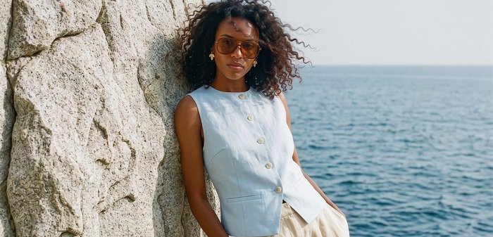 Femme portant un haut sans manches bleu clair avec boutons et des lunettes de soleil, appuyée contre un mur rocheux avec l'océan en arrière-plan.