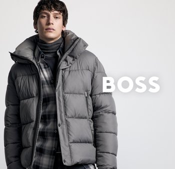 Junger Mann in grauer Pufferjacke, dunklem Rollkragenpullover und kariertem Hemd, vor einem schlichten weißen Hintergrund mit dem Text "BOSS".