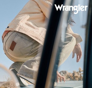 Person som sitter på huk utendørs iført lyse beige jeans og jakke med Wrangler-logo synlig på lommen, klar himmel i bakgrunnen.