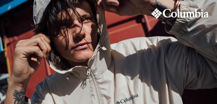 Man with tattoo on wrist adjusting hood of light beige Columbia jacket outdoors in sunlight against red background.