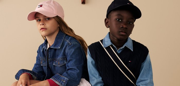 Jeansjacke, rosa Kappe, roter Akzent. Dunkle Weste mit weißem Träger über blauem Hemd. Neutraler Hintergrund. Zwei Kinder nebeneinander sitzend.