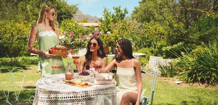 Tres mujeres con gafas de sol disfrutando de bebidas y aperitivos en una mesa redonda de jardín con flores y vegetación de fondo.