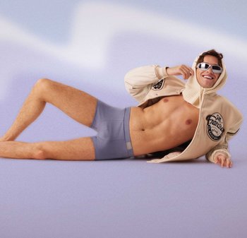 Giacca varsity beige con logo, pantaloncini boxer grigi chiari, texture liscia. Il soggetto è sdraiato su uno sfondo a gradiente viola in posizione informale.