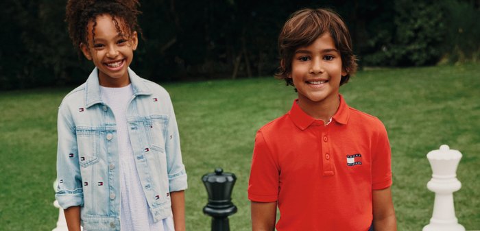 Zwei Kinder lächeln auf einem großen Schachbrett im Freien mit übergroßen schwarzen und weißen Schachfiguren auf dem Gras.