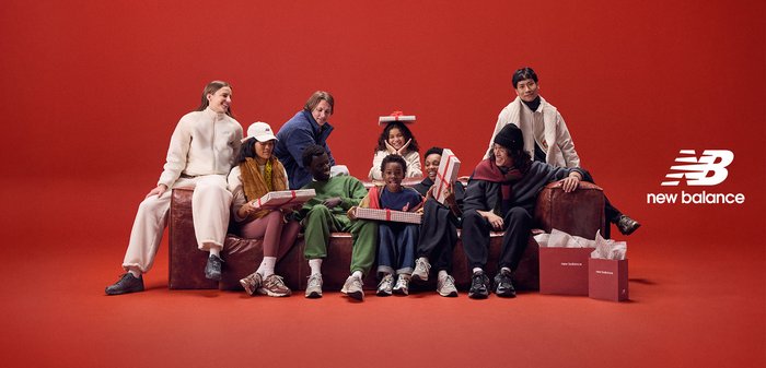 Grupo de personas diversas sentadas en un sofá marrón, vistiendo varios atuendos casuales de telas suaves, con regalos y un fondo rojo.