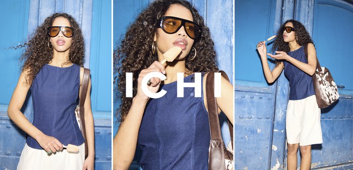 Frau mit lockigem Haar, die eine Sonnenbrille und ein blaues Oberteil trägt, isst ein Eis am Stiel, während sie an einer blauen Metalltür steht.