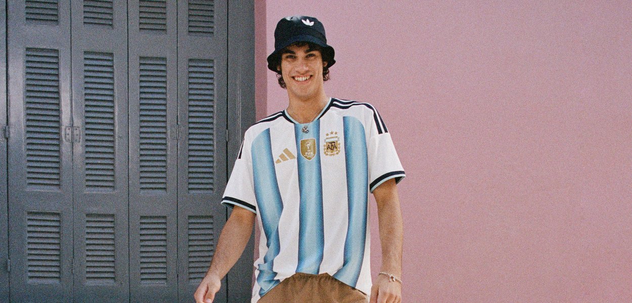 Jeune homme souriant, portant un maillot de football d'Argentine, chapeau seau noir, debout devant un mur rose avec des volets gris fermés.