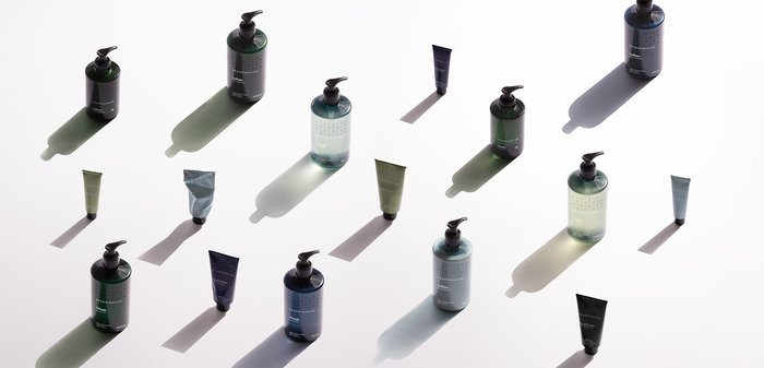 Trois grandes bouteilles avec des bouchons et des étiquettes noirs et verts, ainsi que quatre tubes de teintes vertes variées, projettent de douces ombres sur une surface blanche.