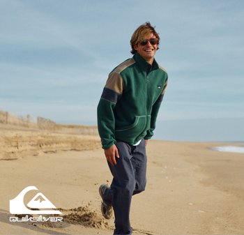 Veste en polaire verte à fermeture éclair avec des accents beiges et marine, poches avant, et pantalon marine décontracté, sur fond de plage.