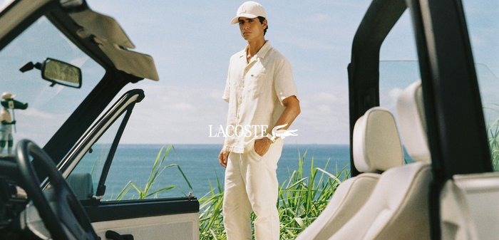 Hombre con ropa informal blanca y gorra junto a la puerta abierta de un coche descapotable cerca de vegetación costera con el océano al fondo.
