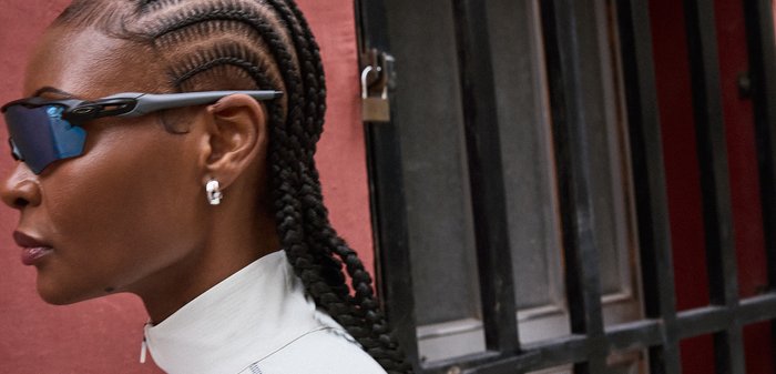 Άτομο με πλεγμένα μαλλιά, που φοράει σκούρα γυαλιά ηλίου τύπου wraparound και μικρούς κρίκους σκουλαρίκια, στέκεται κοντά σε μαύρα μεταλλικά κάγκελα και έναν κόκκινο τοίχο.
