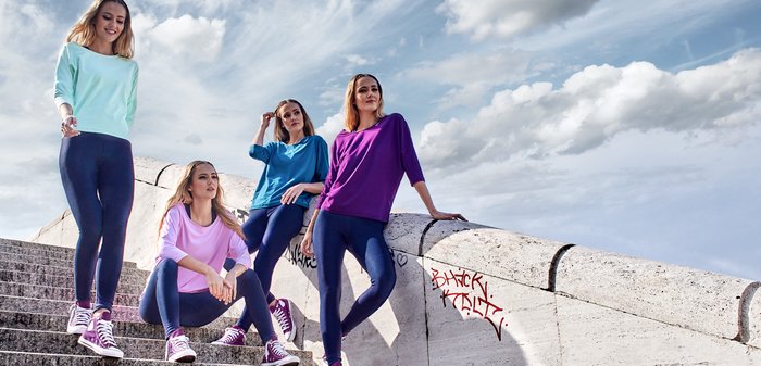 Vier vrouwen dragen kleurrijke, lichte longsleeves (mintgroen, blauw, roze, paars) met donkergekleurde leggings en paarse sneakers, poseren op traptreden.