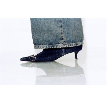 Piede con scarpa a punta in denim scuro con tacco kitten e fibbia argento, abbinata a jeans azzurri sbiaditi che coprono la caviglia.