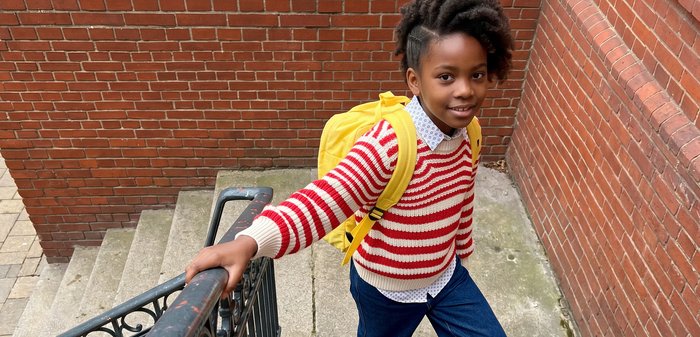 Jeune fille avec un sac à dos jaune montant des escaliers extérieurs à côté d'un mur en briques, tenant la rampe et regardant la caméra.